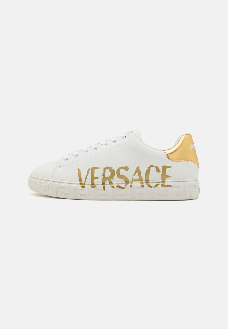 White leather sneaker with gold accents, featuring "VERSACE" logo in bold lettering, and a patterned white sole with geometric detail.