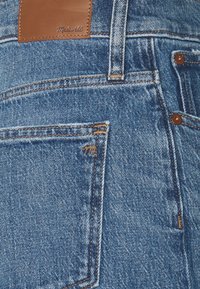 Detailní záběr na zadní kapsu modrých denimových džín s hnědou koženou etiketou Madewell a měděnými nýty na švech.