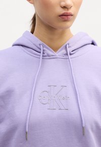 Világoslila kapucnis felső, húzózsinórral, az elején ezüst Calvin Klein logóval. Puha anyag, sima textúrával.