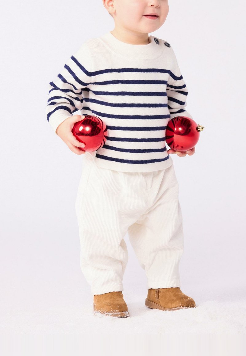 Maglione a righe blu navy e crema, pantaloni di corduroy bianchi, stivali marroni; bambino che tiene due ornamenti rossi. Sfondo bianco con una texture simile alla neve.
