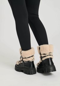 Botas de cuero negras con suelas gruesas, forro y puños de felpa crema, y cordones decorativos. Llevadas con leggings negros.
