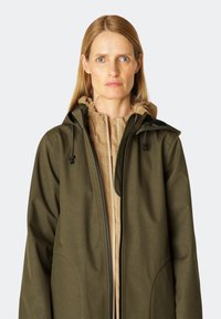 Olijfgroene jas van textuurstof met een lichtgewicht beige binnenlaag. Heeft een hoge kraag en verstelbare trekkoorden op de capuchon.