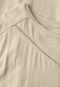 T-shirt en tissu beige clair avec un col rond et un panneau texturé orné de broderies en œillet le long de la couture de l'épaule.