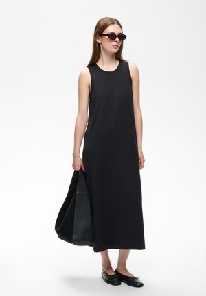 Mujer con vestido midi negro sin mangas, gafas de sol negras y zapatos planos negros que lleva un gran bolso tote negro, de pie frente a un fondo sencillo.