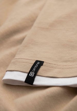 Tessuto beige con orlo cucito e un'etichetta rettangolare nera con la scritta "ombre" su un tessuto bianco sovrapposto sottostante.