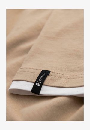 Beige stof met gestikte zoom en een zwart rechthoekig label met de tekst "ombre" op een gelaagde witte stof eronder.