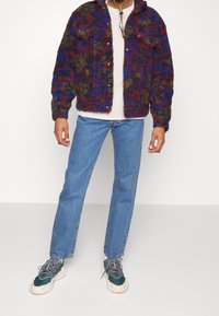 Puszysta, wzorzysta kurtka w ciemnofioletowych, zielonych i czerwonych odcieniach, zapinana na guziki, z dwiema kieszeniami na piersi, noszona na jasnobeżowej koszuli i niebieskich dżinsach.