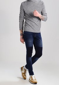 Grauer strukturierter Rollkragenpullover, dunkelblaue Jeans mit hellblauen Flecken und gelbe Sneakers mit weißen Akzenten. Tattoo am Arm sichtbar.