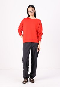 Red oversized sweatshirt with long sleeves paired with loose black trousers and leopard print shoes. Plain background.