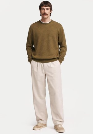 Homme debout les mains dans les poches, portant un pull vert olive, un pantalon blanc et des baskets beiges sur un fond blanc uni.