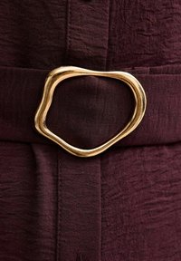 Tessuto bordeaux con finitura testurizzata presenta un fibbia ondulata color oro su una cintura. La fibbia è asimmetrica e dal design liscio.