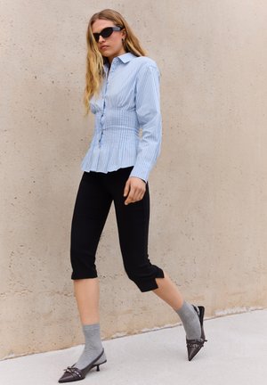 Femeie care merge pe lângă un perete bej, purtând o cămașă albastră cu dungi, pantaloni negri scurți, șosete gri, pantofi negri cu toc ascuțit și bretele și ochelari de soare negri.