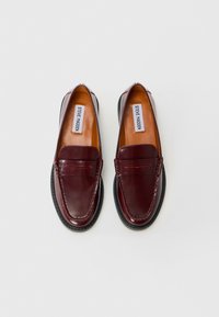 Paire de mocassins en cuir marron foncé avec semelles noires et étiquette « Steve Madden » à l'intérieur, présentée sur un fond clair uni.