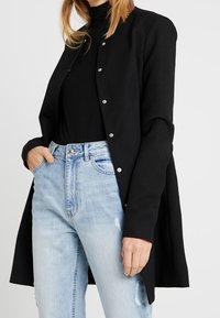 Cárdigan negro con un tejido texturizado, cierre de botones a presión, mangas largas y un diseño ligeramente acampanado, llevado sobre un cuello de tortuga negro y unos vaqueros azul claro.