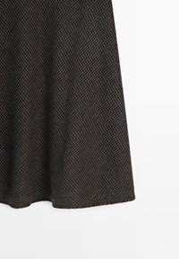 Tissu gris foncé avec un petit motif à carreaux noirs tombant vers le bas, montrant la partie inférieure d’un vêtement sur fond blanc.