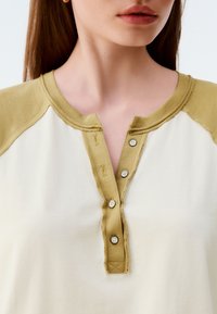 Long-sleeve top with a cream body and olive green raglan sleeves. Features a buttoned henley neckline with four buttons and visible stitching.