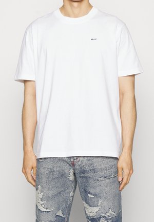 Biała bawełniana koszulka z okrągłym dekoltem, krótkimi rękawami oraz małą czarną nadrukowaną etykietą "NN. 07" na lewej piersi. Noszona z podartymi dżinsami.