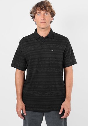 Homme aux cheveux bruns bouclés portant un polo noir à manches courtes avec un motif et un pantalon gris, debout devant un fond clair uni.