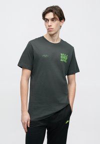 T-shirt de algodão cinzento escuro com um design gráfico verde néon no peito, mangas curtas e decote redondo, usada com calças pretas.