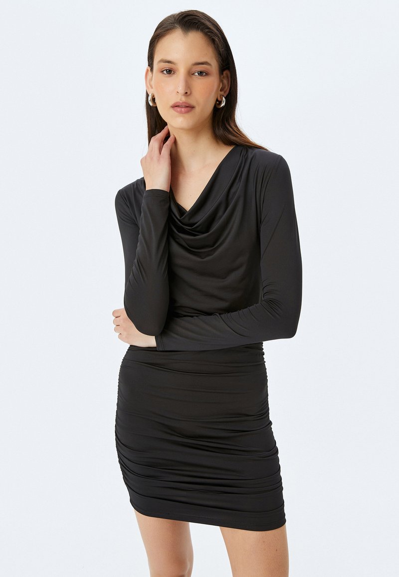 Musta pitkähihainemekko, jossa on laskostettu kaula-aukko ja rypytetty yksityiskohta. Valmistettu sileästä, joustavasta kankaasta, joka istuu tiukasti vyötäröllä ja lantiossa.