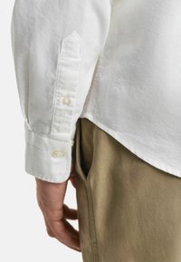 Weißes Langarmhemd mit geknöpften Manschetten, aus strukturiertem Stoff. Kombiniert mit hellen Khakihosen, die eine Seitenansicht der Tasche zeigen.