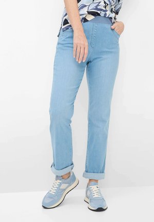 Person iført lyseblå op rullede jeans og matchende blå sneakers, står med den ene hånd i lommen mod en hvid baggrund.