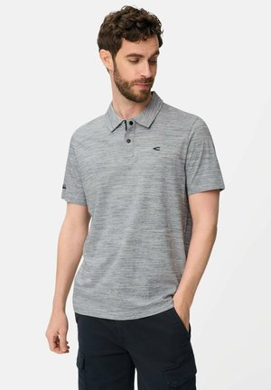 Mand iført en kortærmet grå polo og sorte cargobukser, stående med den ene hånd i lommen, hovedet bøjet ned og til siden.