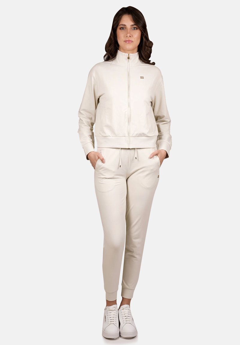 Giacca zip-up beige chiaro con collo alto e polsini a costine, abbinata a pantaloni jogger affusolati abbinati e sneaker bianche. Tessuto morbido, design minimale.