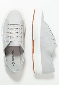 Les baskets en toile grise présentent une tige texturée, une semelle en caoutchouc blanche et un marquage discret. Le design comprend six œillets pour les lacets.