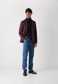 Burgundowy blezer na ciemnym golfie, niebieskie dżinsy i czarne buty; model w czapce na czarno, stoi na neutralnym tle.