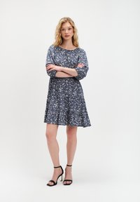 Granatowa sukienka w kwiaty z rękawami o długości trzech �ćwierci, marszczonym pasem i rozkloszowanym dołem, noszona z czarnymi sandałami na obcasie. Lekki wzór kwiatowy na całej powierzchni.