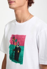 Hvid bomulds T-shirt med et farverigt, abstrakt grafisk design med en figur i sort tøj og et gyldent ansigt, suppleret med livlige baggrundsfarver.
