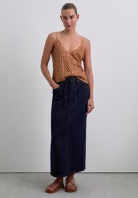 Top sin mangas de bronce con un patrón texturizado, combinado con una falda larga de denim oscuro con cintura ajustable y bolsillos laterales. Sandalias marrones.