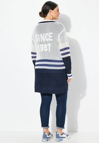Lang, åbent strikket kardigan i hvid med marineblå striber, prydet med "SINCE 1987" på ryggen, parret med mørke jeans og blå sneakers.