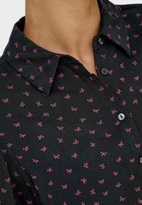 Gros plan sur une chemise noire à boutons avec de petits motifs de nœud rose et un col pointu sur le cou et le haut de la poitrine d'une personne.