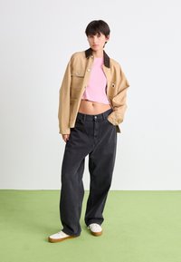 Veste beige cropped avec col marron, haut rose côtelé, jean noir ample et baskets blanches avec semelles marron sur un sol vert.