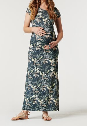 Zwangere vrouw die een korte mouw, bloemenjurk tot op de grond en sandalen draagt, met beide handen voorzichtig haar buik vasthoudend.