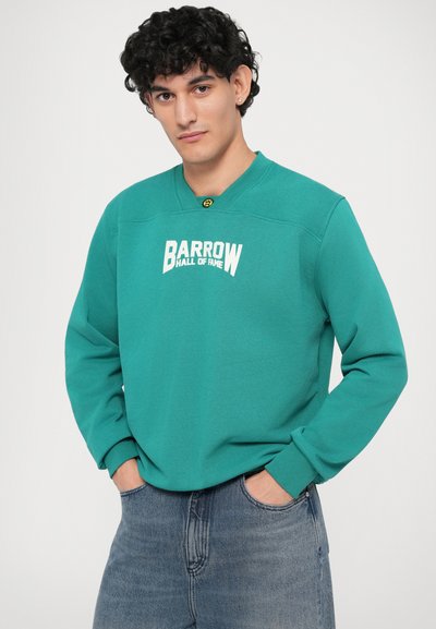 Felpa teal con colletto a crew, con la scritta bianca "BARROW HALL OF FAME" sul davanti, abbinata a jeans di denim blu chiaro.