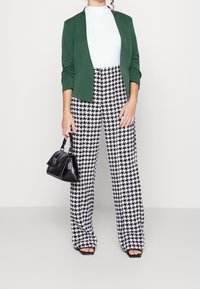 Zelený blazer, bílý rolák, kalhoty v černo-bílém houndstooth vzoru, černá kabelka a černé sandály s otevřenou špičkou. Formální outfit s přiléhavými a volnými prvky.