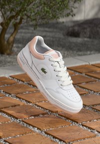 Fehér bőr sneaker rózsaszín akcentusokkal és fehér fűzőkkel. Jellemzője a perforált orr, texturált gumi talp, és a Lacoste embléma az oldalon.
