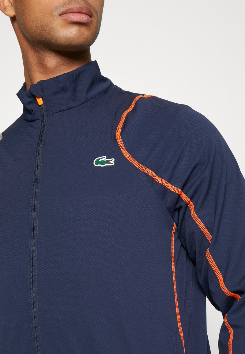 Marineblauwe jas met hoge kraag, oranje stiksels en een groen Lacoste-logo. Voorzien van een ritssluiting en textiel met structuur.