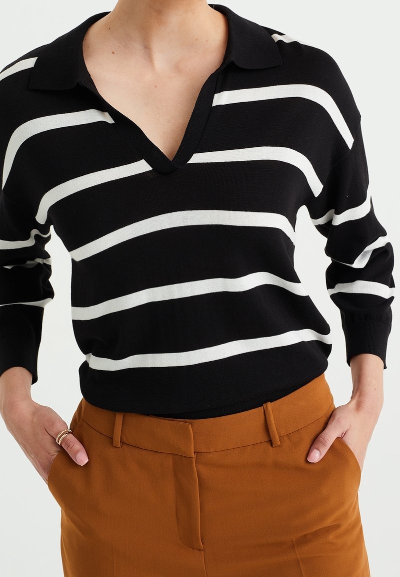 Černobíle pruhovaný svetr s výstřihem do V a límcem, s dlouhými rukávy. Nosený s hnědými, přiléhavými kalhotami s vysokým pasem.