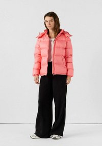 Giacca a vento rosa con chiusura zip, design trapuntato, cappuccio e polsini elasticizzati, abbinata a pantaloni neri larghi e sneakers argentati.
