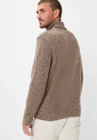 Brauner Strickpullover mit hohem Kragen, strukturiertem Stoff und blauen Sprenkeln. Das Model steht mit teilweise zur Seite gedrehtem Rücken und trägt helle Hosen.