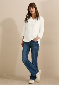 Blusa blanca texturizada con cuello, combinada con jeans de pierna recta azules y zapatillas blancas. La parte superior tiene mangas de 3/4 y un corte relajado.