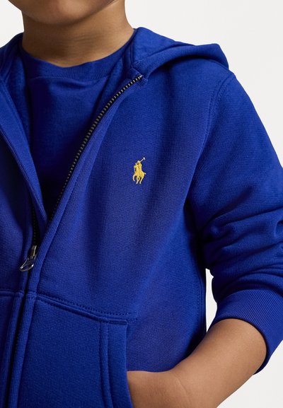 Μπλε φούτερ με φερμουάρ, κατασκευασμένο από μαλακό υλικό, με μικρό κίτρινο λογότυπο στο αριστερό στήθος και τσέπη καγκουρό στη μέση.