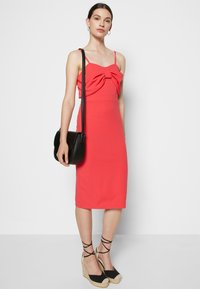 Vestido justo coral com alças finas, detalhe em nó na zona do busto, combinado com uma mala preta de ombro e plataformas pretas em espadrille.