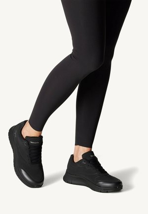 Osoba ubrana w czarne legginsy i czarne sneakersy z grubą podeszwą stoi na jasnym tle.