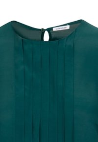 Turkusowa bluzka z pionowymi fa�łdami, okrągłym dekoltem i zapięciem na guziki z tyłu. Materiał wydaje się gładki z delikatnym połyskiem. Widoczna etykieta marki.