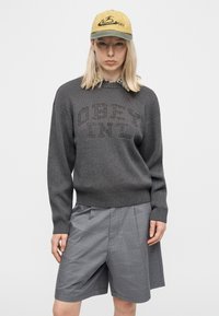 Άτομο που φοράει γκρι πουλόβερ με το κείμενο "OBEY INT.", γκρι καρό σορτς, κίτρινο και πράσινο καπέλο με σχέδιο φιδιού, στέκεται μπροστά σε λευκό φόντο.